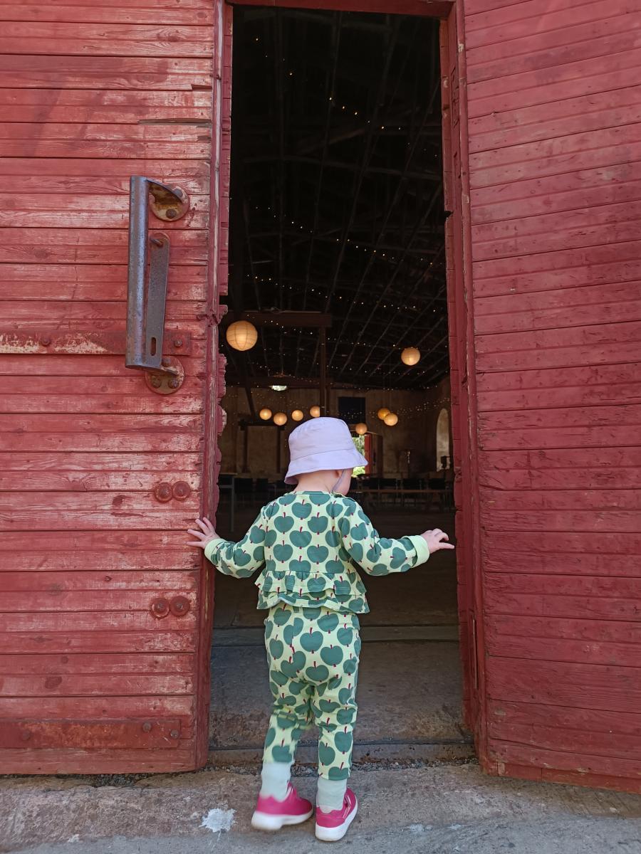 Lapsi vihreissä vaatteissa kurkkaa vanhan tehdashallin punaisesta ovesta sisään.